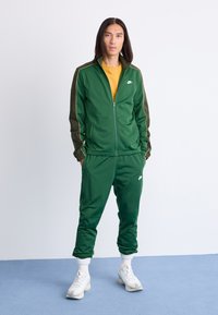 Survêtement Nike vert avec veste zippée et pantalon à taille élastique, complété par une chemise jaune. Comprend un logo blanc et des accents contrastés.