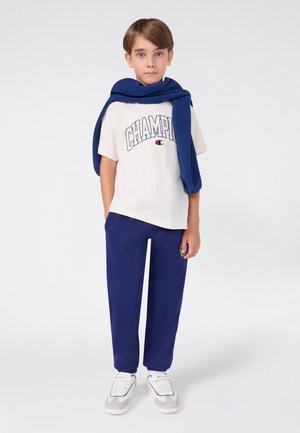 Junge steht mit Blick nach vorne, trägt ein weißes Champion-T-Shirt, eine blaue Jogginghose, weiße Sneakers und einen blauen Hoodie, der über den Schultern liegt.