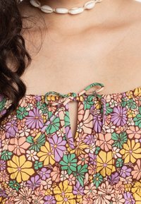 Gros plan sur l'épaule et la poitrine portant une robe fleurie avec des fleurs vertes, violettes et jaunes, ornée d'un col à nœud, associée à un collier de coquillages blanc.