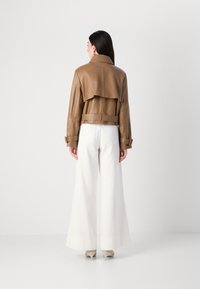 Een vrouw staat met haar rug naar de camera, gekleed in een bruine leren cropped jack en witte wijde broek, met een minimalistische stijl.