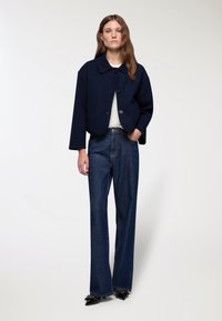 Giacca corta blu scuro con colletto e tre bottoni, abbinata a jeans scuri a vita alta e scarpe nere a punta.