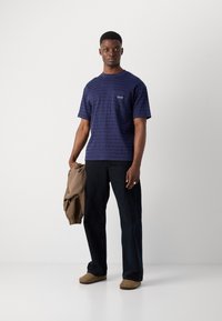 T-shirt bleu marine à manches courtes avec un motif texturé, doté d'un petit logo sur la poitrine gauche. Pantalons foncés et pantoufles beiges avec une veste.