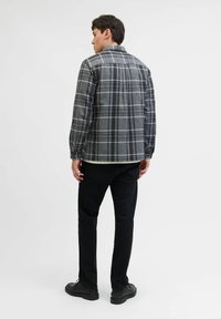 Camisa chaqueta a cuadros grises con mangas largas, cuello y vaqueros negros. El modelo está de pie un poco de lado; textura suave y corte relajado.