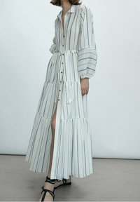 Robe longue à rayures blanches, avec un devant boutonné, des manches évasées, un design à étages et une fente sur le côté. Le matériau semble léger et aérien.