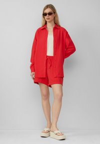 Czerwona, teksturowana koszula z guzikami, pasujące czerwone szorty i off-white sandały na platformie. Modelka nosi ciemne okulary przeciwsłoneczne. Prosty, casualowy strój.