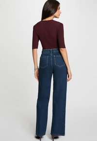 Frau, die sich abkehrt, trägt ein enges weinrotfarbenes Oberteil mit Ellenbogenärmeln sowie eine hoch taillierte, weite blaue Jeans und Absatzschuhe.