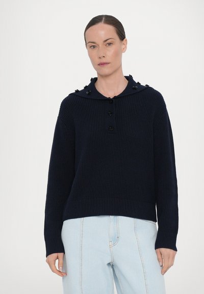 Marinblå stickad tröja med stor krage, utrustad med knappstängning och dekorativa detaljer på axlarna, i kombination med ljusblå jeans.