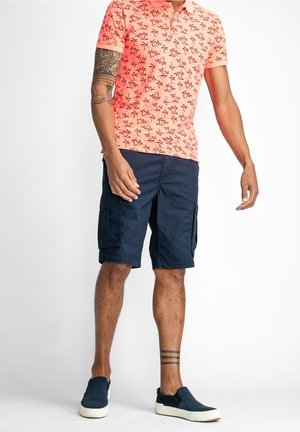 Polo en corail avec motif d'arbres tropicaux noirs, associé à un short cargo bleu marine. Modèle portant des chaussures slipper bleu marine, affichant des tatouages sur les bras.