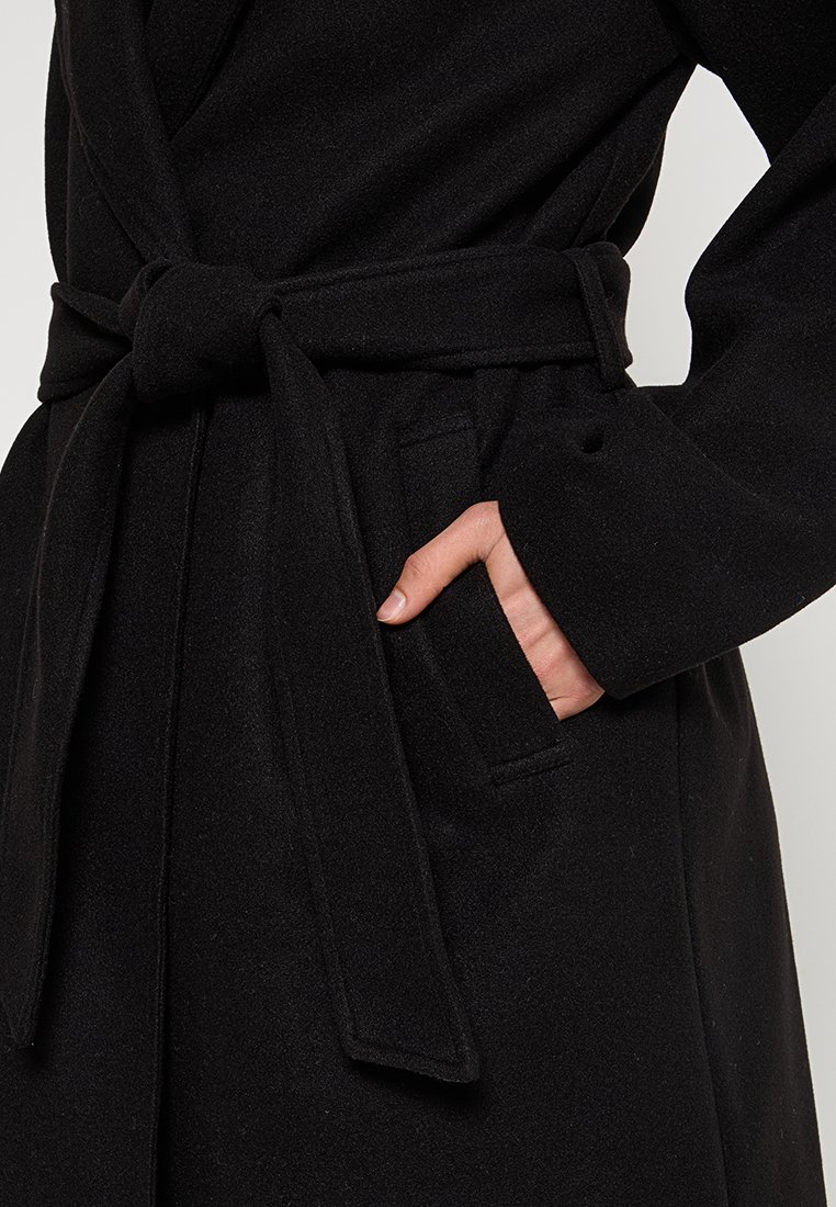 Svart ullkappa med en strukturerad yta, utrustad med ett knytbälte i midjan och sidofickor, visad från midjan och uppåt, som framhäver designens detaljer.