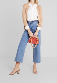 Beżowy kardigan, biała koszula, jasnoniebieskie jeansy z szerokimi nogawkami i wysokim stanem oraz beżowe sandały na słupku. Czerwona torebka o fakturze z łańcuszkowym paskiem.