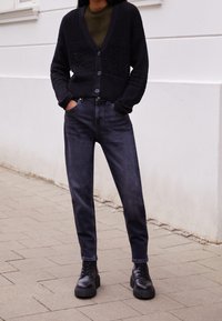 Cárdigan negro de punto con patrones texturizados, botones, combinado con jeans oscuros y botas negras de suela gruesa sobre una superficie pavimentada.