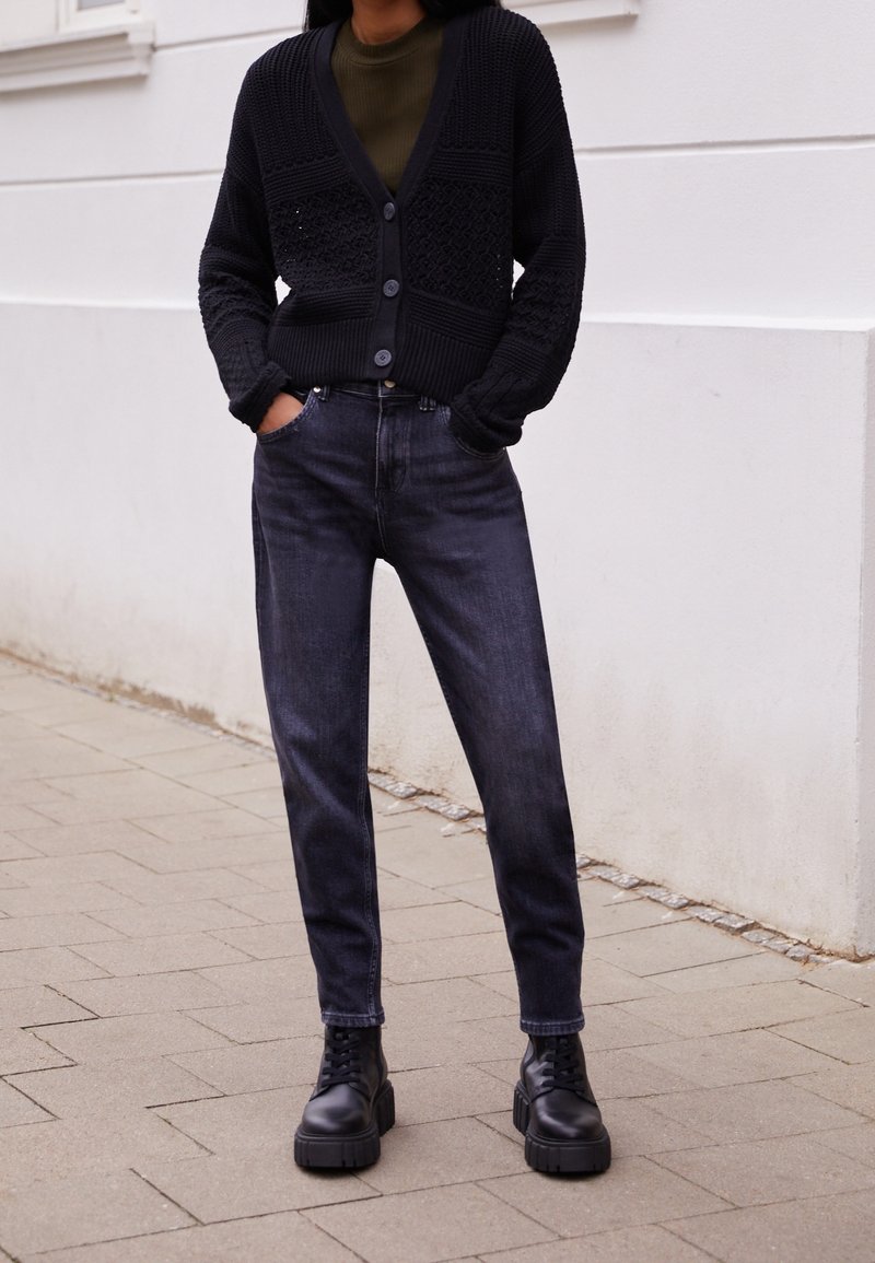 Cárdigan negro de punto con patrones texturizados, botones, combinado con jeans oscuros y botas negras de suela gruesa sobre una superficie pavimentada.