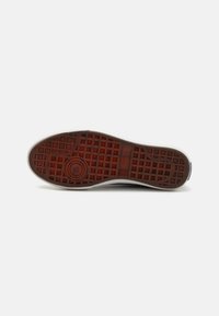 Suela de zapatilla de goma con un patrón de cuadrícula texturizado en rojo y negro, con detalles circulares. Bordes blancos y un acabado suave alrededor del perímetro.
