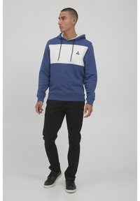 Felpa con cappuccio blu e bianca con logo triangolare, caratterizzata da un frontale a blocchi di colore e cordini regolabili. Abbinata a pantaloni e scarpe neri.