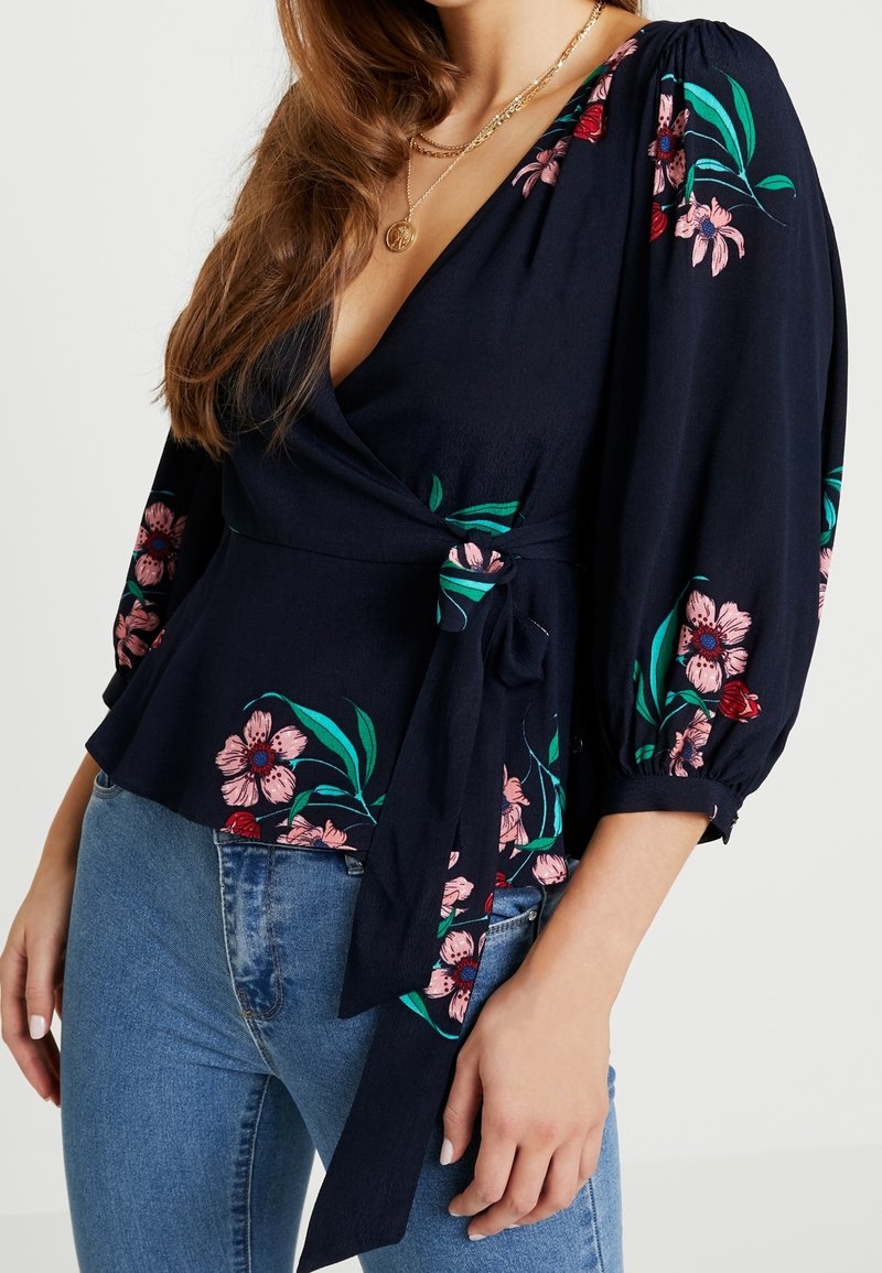 Blusa avvolgente blu navy con stampa floreale in rosa e rosso. Presenta maniche a sbuffo e vita legata. Abbinata a jeans azzurri chiari.
