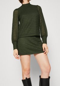 Grön texturerad stickad tröja med långa puffärmar, matchad med en mini-kjol. Båda har ett enhetligt diamantsmönster. Svarta knähöga stövlar.