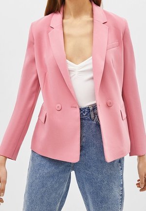 Woman wearing a fitted pink blazer over a white ruched top and high-waisted blue jeans, standing against a plain background.