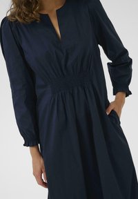 Dunkelblaues Kleid mit geraffter Taille, langen Ärmeln und einem Schlüssellochausschnitt, das eine entspannte Silhouette und strukturierten Stoff aufweist.