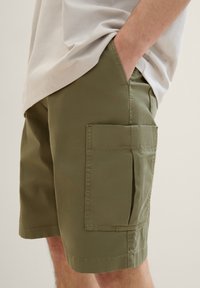Oliwkowe szorty cargo wykonane z lekkiego materiału; mają dużą boczną kieszeń oraz gładką, równą strukturę z prostym wykończeniem.