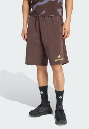 Person trägt braune Sportschuhe mit gelbem Logo, schwarze Kniestrümpfe mit weißen Logos und schwarze Laufschuhe und steht auf weißem Boden.