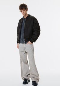 Ung mand iført en sort bomberjakke, navy polo-sweater, lysegrå brede jeans med sidelæderstriber og sorte sko, stående mod baggrund i hvid.