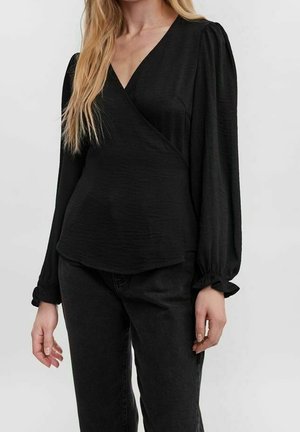 Kobieta w czarnej, długorękawowej bluzce typu wrap z bufkami na ramionach i falbanami na mankietach, w połączeniu z czarnymi spodniami, na jednolitym tle.