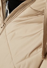 Chaqueta acolchada beige con cierre de cremallera, capucha y cordón de cuerda texturizada. Presenta un sutil acolchado en forma de rombo para un detalle adicional.