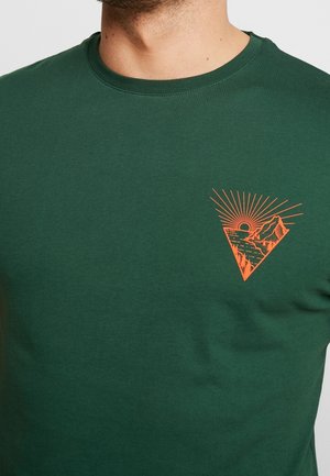 Grünes T-Shirt mit Rundhalsausschnitt und kleinem orangefarbenem Grafikmotiv einer Sonne, die hinter Bergen und einem See auf der linken Brustseite untergeht.