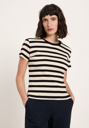 Frau mit schulterlangem dunklem Haar, trägt ein schwarz-weiß gestreiftes kurzärmeliges Oberteil und dunkle Hosen, Hände in den Taschen, vor einem schlichten Hintergrund.