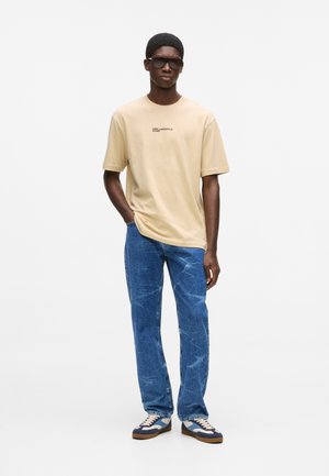 Mand iført beige Karl Lagerfeld Jeans t-shirt, blå jeans, blå og hvide sneakers samt sorte solbriller, stående med den ene hånd i lommen.