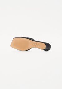 Sandale à talons hauts noire avec une bride texturée, un bout carré et une semelle en caoutchouc beige avec un motif strié. Logo de la marque visible.