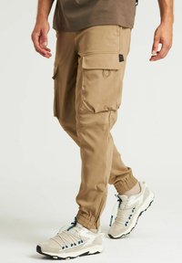 Beige cargobroeken met elastische manchetten, zijzakken en een lusdetail. Draag ze met lichte sneakers met grijze en teal accenten.