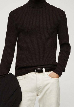 Personne portant un pull à col roulé côtelé noir rentré dans un pantalon blanc avec une ceinture noire, main dans la poche, tenant une veste noire.