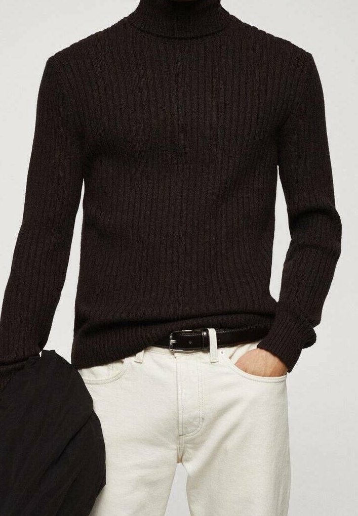 Personne portant un pull à col roulé côtelé noir rentré dans un pantalon blanc avec une ceinture noire, main dans la poche, tenant une veste noire.