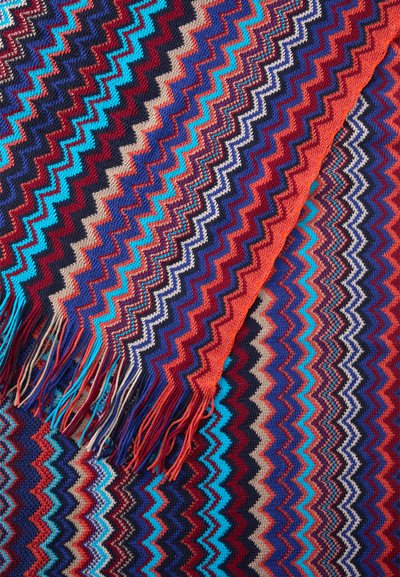 Flerfarvet zigzag mønster tekstil i rød, blå og sort med fransede kanter, der viser en strikket tekstur og levende nuancer.