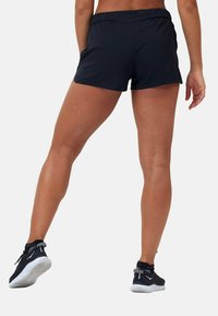 Mustat urheilusortsit, joissa on joustava vyötärö ja jotka korostavat sileää, kevyttä kangasta. Yhdistetty mustiin urheilukenkiin, joissa on valkoinen pohja.