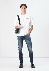 Fehér, rövid ujjú póló piros logóval, kék skinny farmernadrág és fekete cipő, egy kis fekete keresztpántos táskával a kezében. Egyszerű háttér.