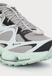 Chaussure de sport en mesh de couleur grise, blanche et noire. Présente une marque Gore-Tex, des accents réfléchissants et une semelle verte menthe. Design supérieur texturé.