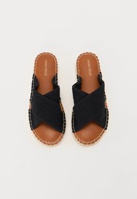 Svart tekstil sandaler med kryssremmer, flate, brune lær såler og beige søm langs kantene. Enkle, åpne tå-design.