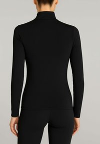 Donna che indossa un top attillato nero a maniche lunghe e pantaloni neri, in piedi con la schiena rivolta verso la macchina fotografica su uno sfondo semplice.