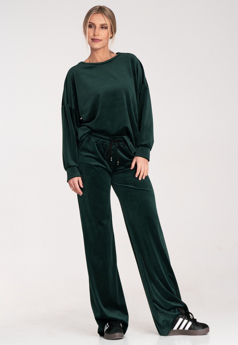 Ciemnozielony, aksamitny zestaw odzieży domowej składa się z luźnej bluzki z opadającymi ramionami oraz spodni z elastycznym pasem. Komplet zestawiony z czarnymi trampeczkami.