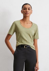 T-shirt a maniche corte con scollatura a barca di colore verde oliva chiaro, realizzata in tessuto morbido, abbinata a pantaloni neri e una cintura con fibbia dorata.