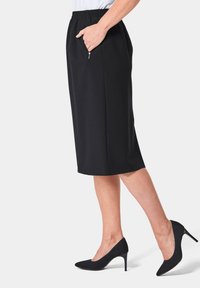 Schwarzer knielanger Rock mit elastischem Bund, seitlichen Taschen und glatter Textur. Kombiniert mit schwarzen High Heels.