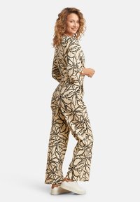Beige jumpsuit met zwart bloemenpatroon, lange mouwen en wijde pijpen. Heeft een relaxed fit en wordt gedragen met witte sneakers.