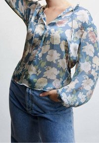 Blomstret bluse med lyseblå baggrund, lange ærmer og V-hals. Stoffet virker gennemsigtigt med farverigt blomsterprint og bånddetaljer.
