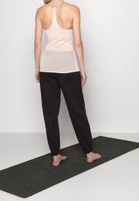 Jasnoróżowy top na ramiączkach w stylu racerback w połączeniu z czarnymi spodniami dresowymi. Top ma dopasowany krój, natomiast spodnie mają elastyczne mankiety. Stoi na macie do jogi.
