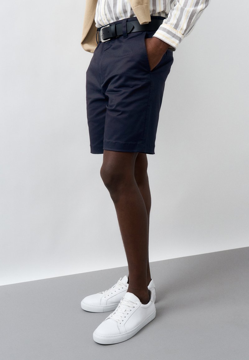 Άνδρας που φοράει σκούρα μπλε σορτς, ριγέ πουκάμισο, μπεζ πουλόβερ περασμένο πάνω από τους ώμους, μαύρη ζώνη και λευκά αθλητικά παπούτσια, στέκεται με τα χέρια στις τσέπες.