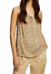 Femme portant un top floral sans manches et ample aux teintes vertes, orange et beige, associé à un pantalon beige, la main dans la poche.