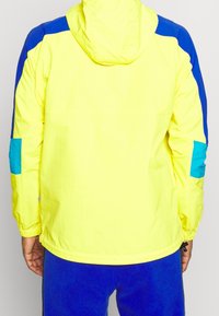 Giacca gialla con cappuccio, caratterizzata da dettagli blu e turchese su spalle e maniche. Realizzata in materiale leggero, con polsini elasticizzati.