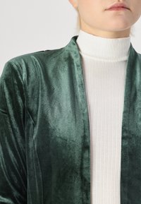 Grön sammetsjacka med mjuk textur, öppen frontdesign. Buren över en vit ribbad polotröja, som visar en slät och kontrasterande stil.